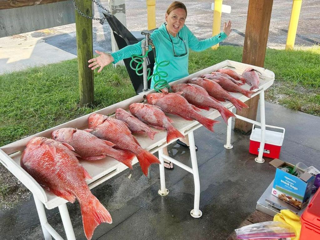 A lady angler stands behind a cleaning table full of all sizes of red snapper.